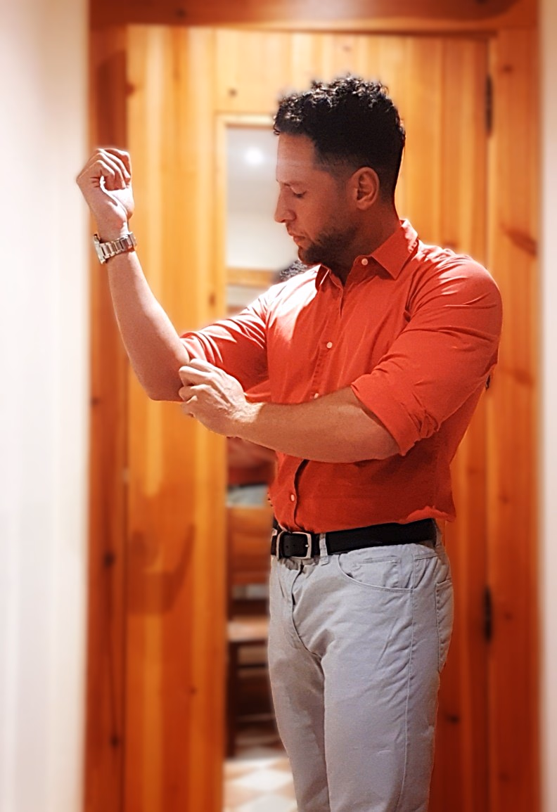 Rolling up the sleeve of a coral shirt in a wood-panelled interior. Alexander, male escort for women and couples.