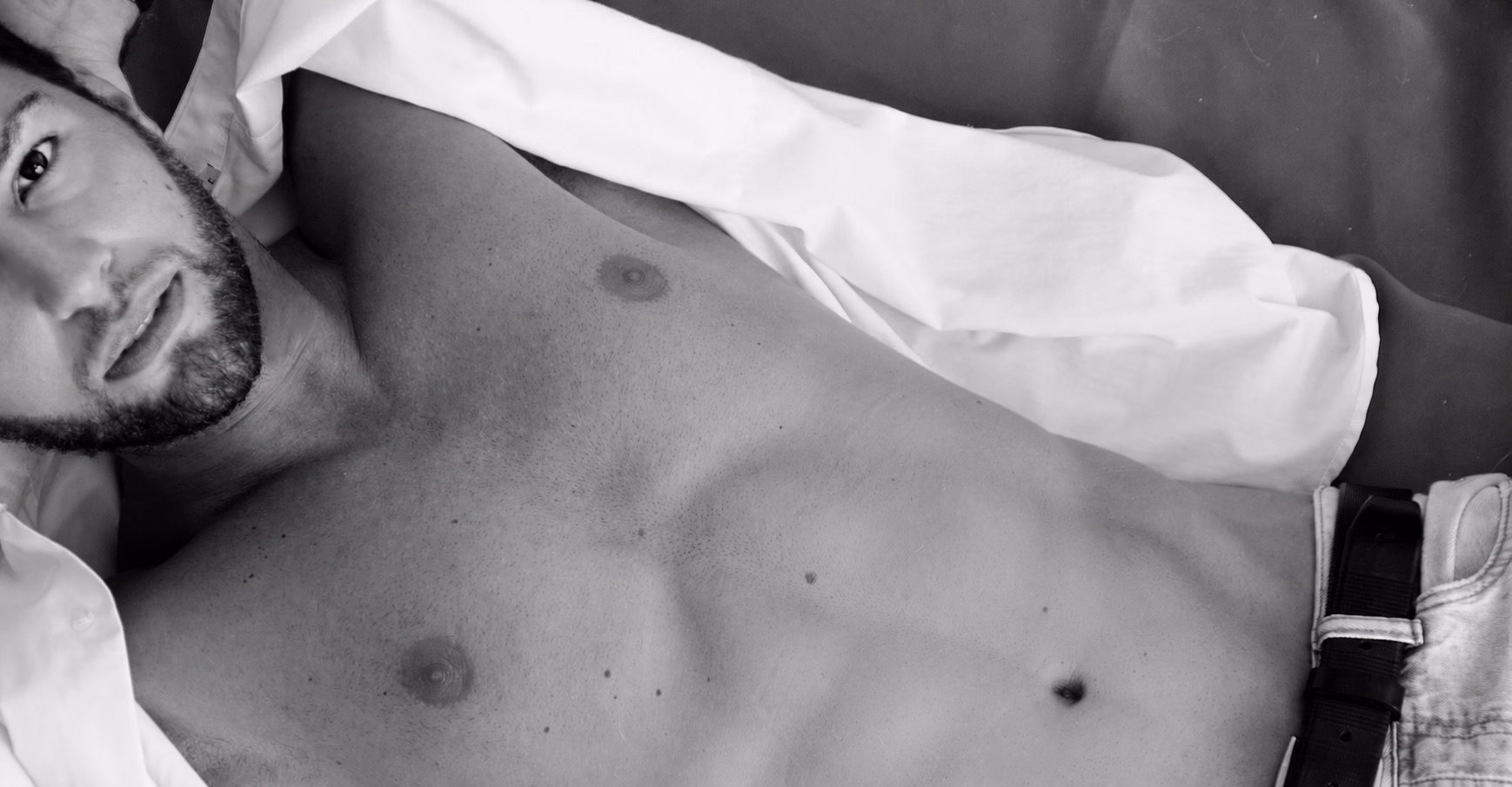 Alexander, male escort, reclining in a black-and-white portrait, shirt open over his chest, gaze to camera.