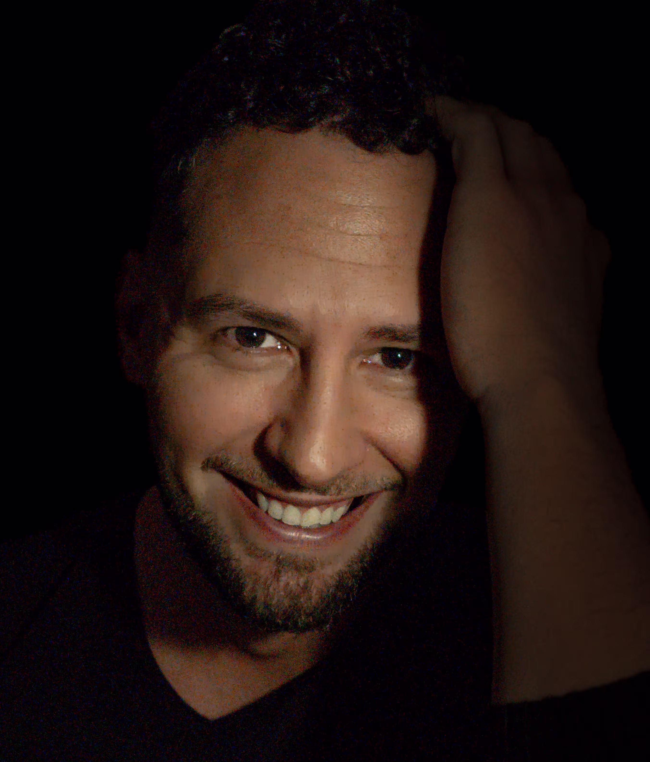 Smiling with a hand in his hair against a warm-toned dark background. A portrait of Alexander, male company for women.