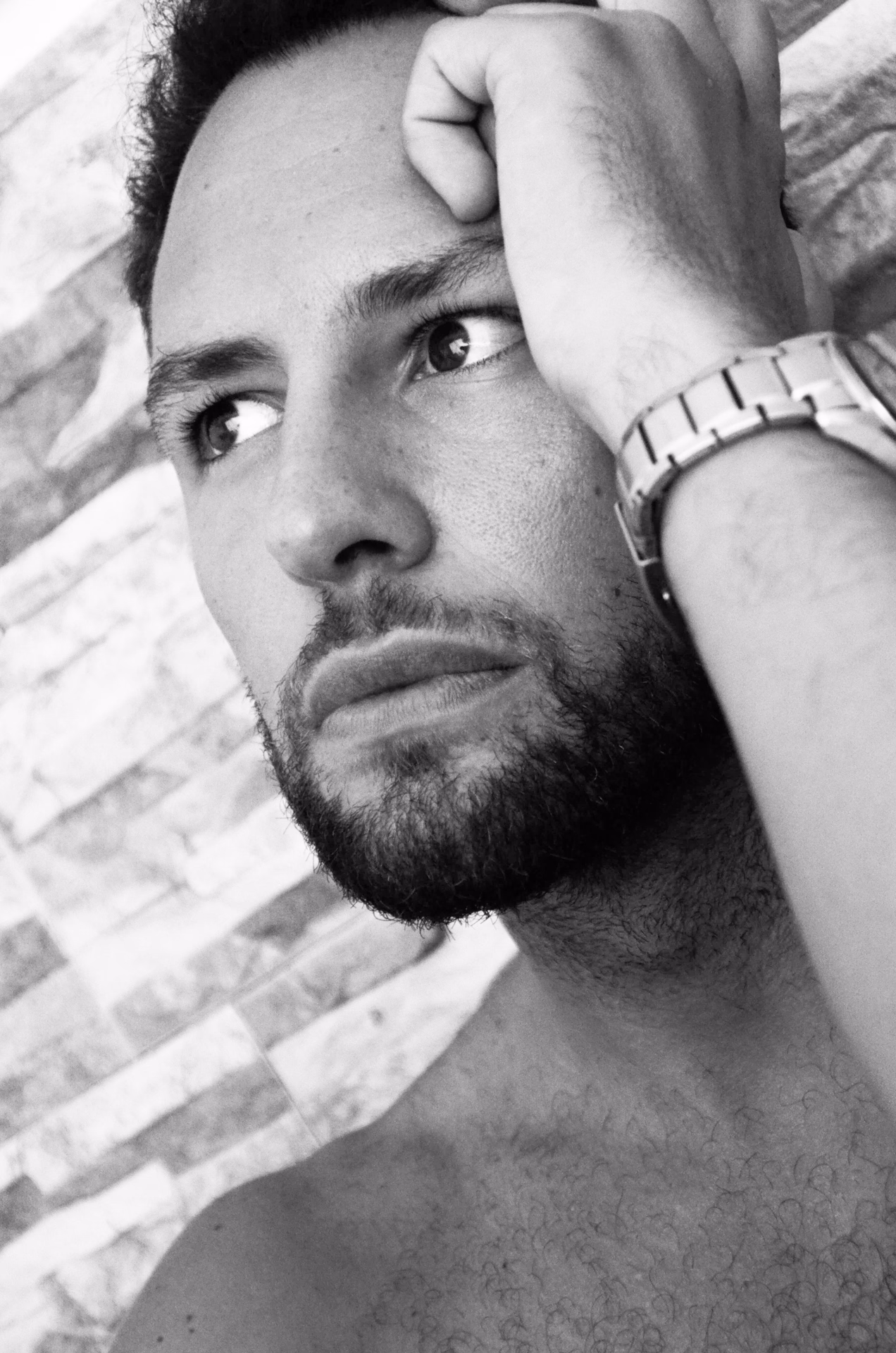 A hand in his hair, a stone wall behind him: a pensive black-and-white portrait of Alexander, escort for women.