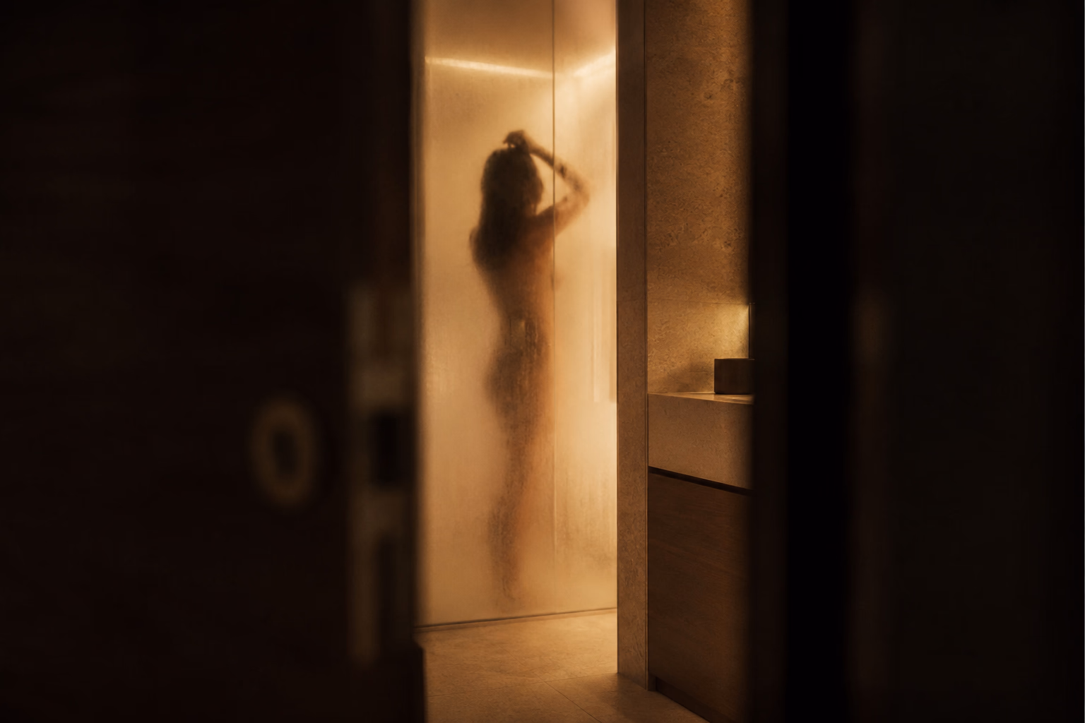 The silhouette of a woman behind the steamed glass of a shower, seen through the gap of a half-open door: in non-consensual voyeurism, the gaze has not been invited, and that is what separates it from cuckolding.
