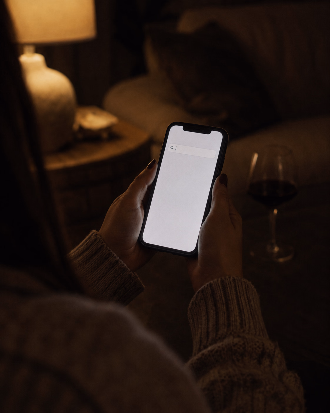 A woman's hands hold a phone in the dim light of a living room, with the search field still empty and the cursor blinking, next to a glass of red wine: the moment when you search for the difference between a gigolo and an escort and do not even know which word to type.
