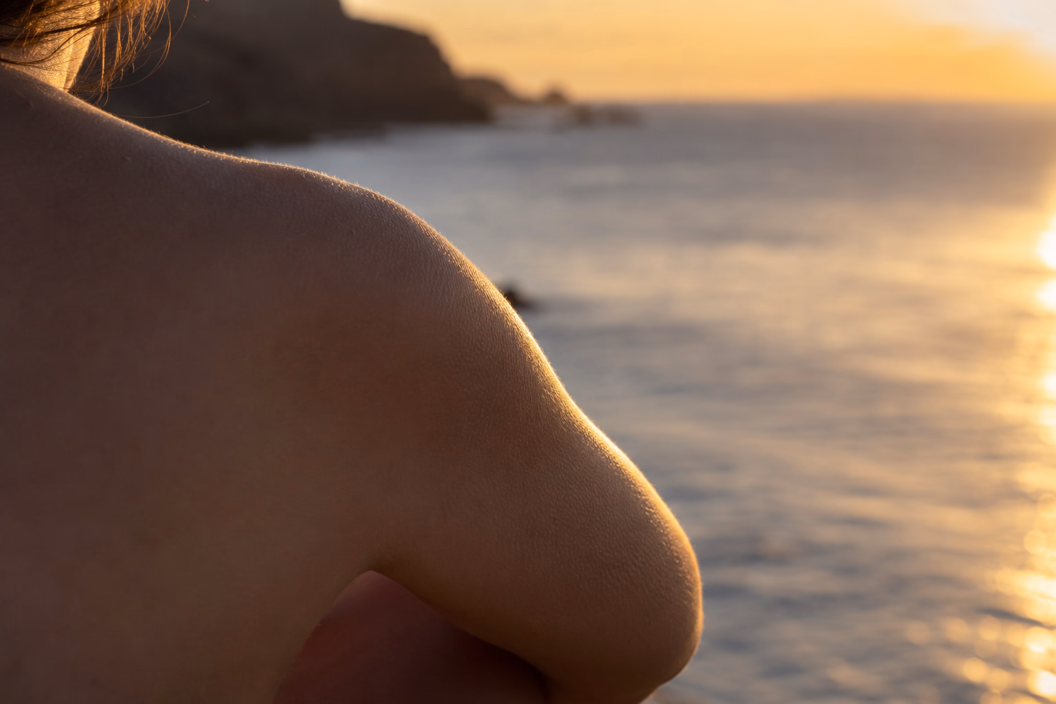 Spalla di una donna in controluce nel tardo pomeriggio, con il sole basso sull'Atlantico e la costa delle Canarie sfocata sullo sfondo.