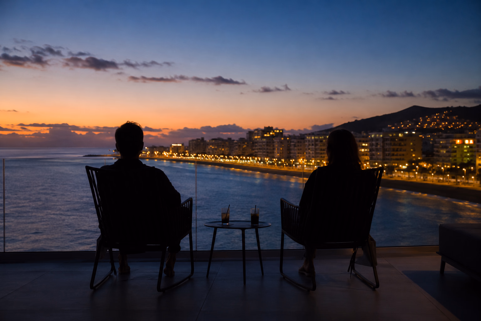 Due sagome sedute su una terrazza sul mare all'ora blu, una donna e il suo escort, con due bicchieri sul tavolo e le luci della costa che si accendono.