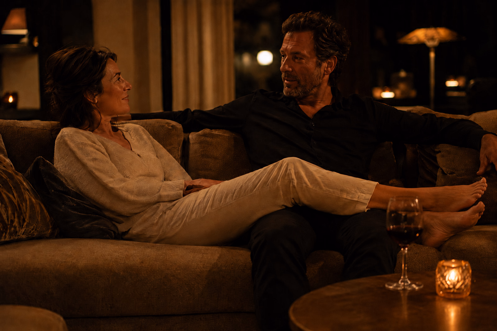 A middle-aged couple talking, relaxed on a sofa one night, in low light; she is reclining with her legs across his lap, a glass of wine and a candle on the low table.