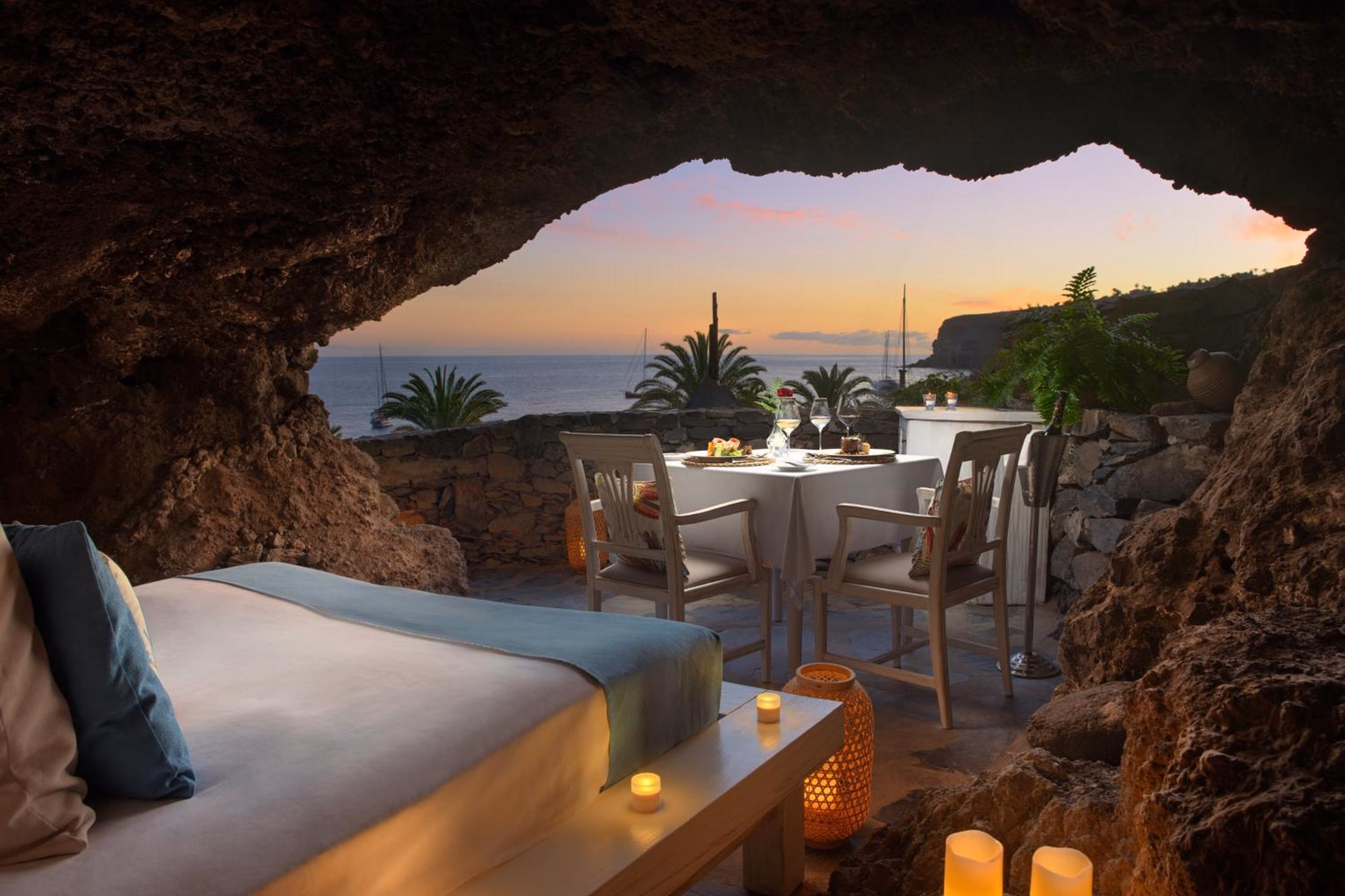 Ein Tisch und ein Bett in einer Grotte an der Klippe, mit Kerzen und dem Meer bei Sonnenuntergang