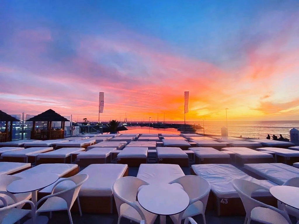 I letti bianchi del Kaluna Beach Club al tramonto, con la piscina a sfioro e il porto di Puerto Colón sullo sfondo